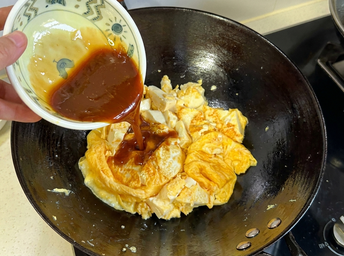 小さなボウルから濃い茶色のタレを、中華鍋の中の卵と豆腐のミックスに注ぎ入れている様子。