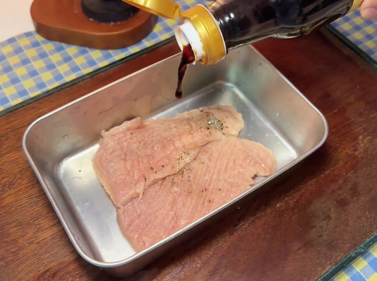 金属製のバットに入った味付きの豚肉に、ガラス瓶から濃口醤油を注いでいる様子。