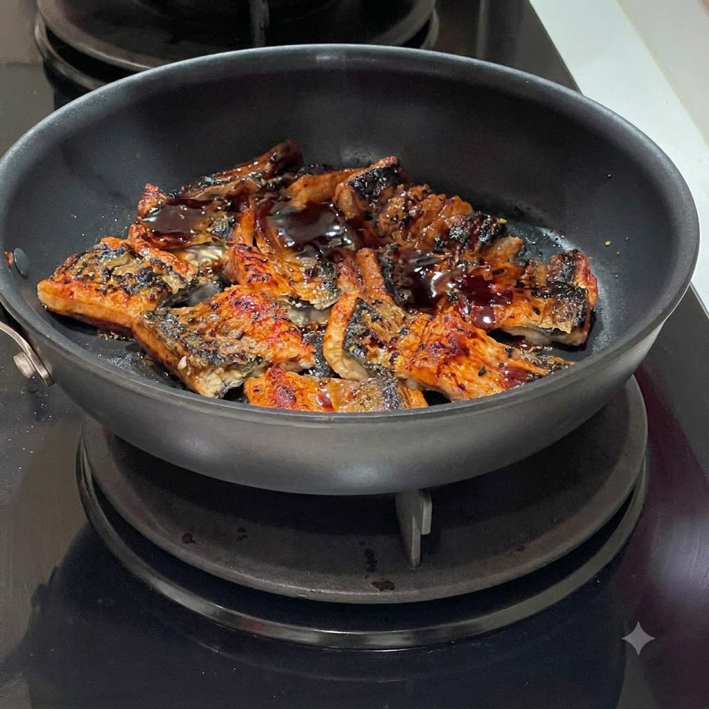 フライパンの中でジューシーに焼けているうなぎの切り身に、濃い色のタレを回しかけている様子。