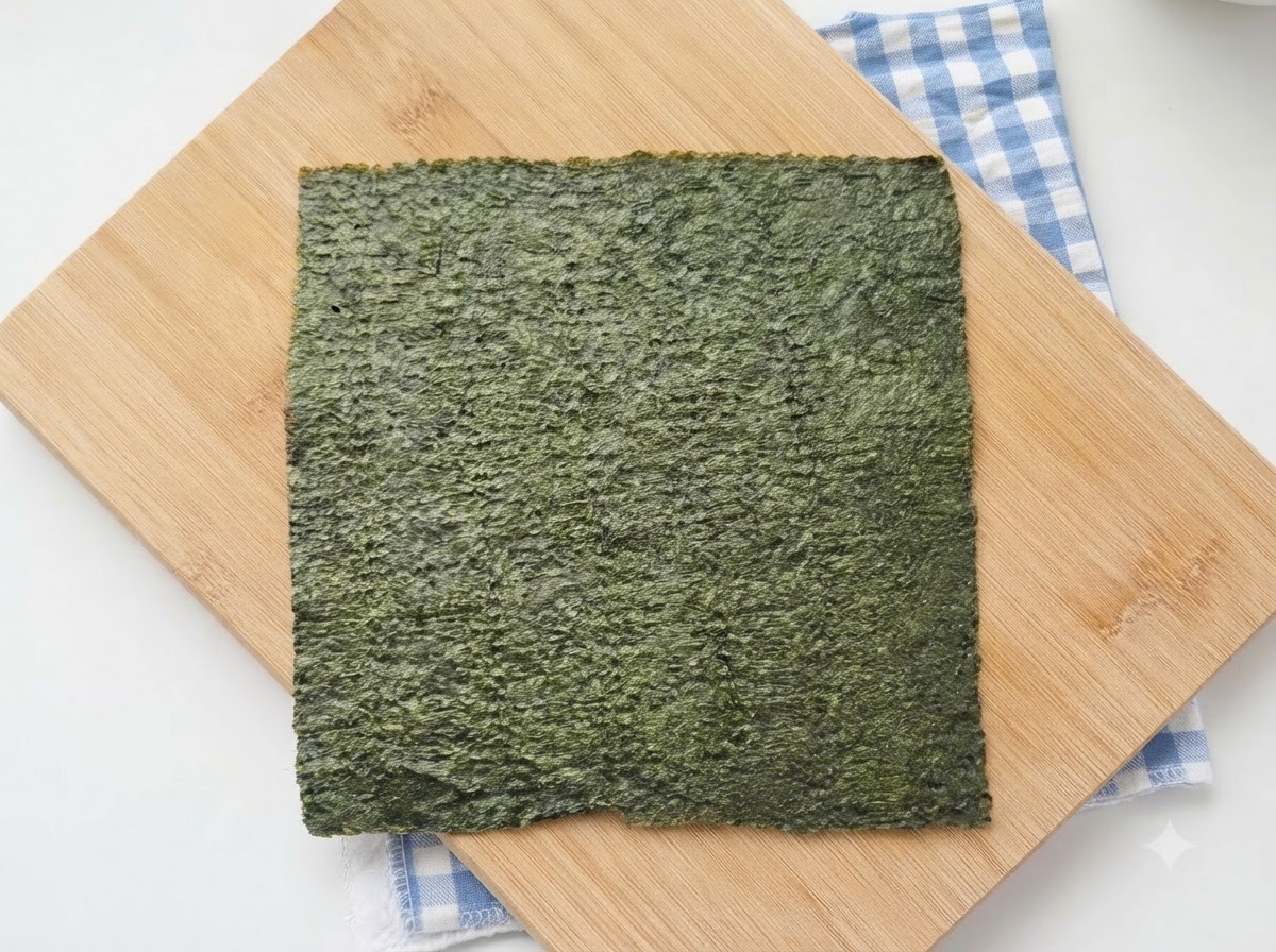青いチェック柄の布を敷いた木製のまな板の上に、緑色の寿司海苔の全形が置かれている様子。