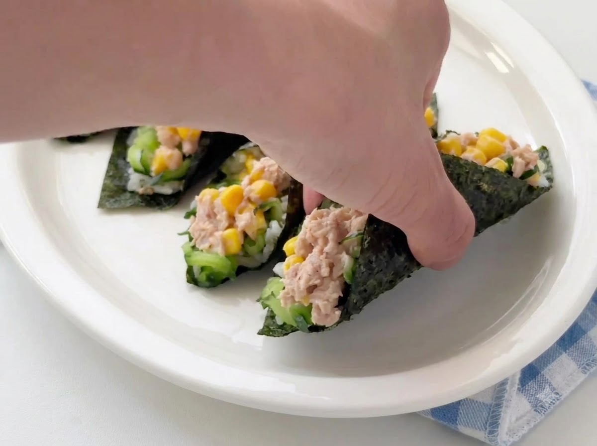 白いサービングプレートの上で、完成したツナと海苔のご飯ロールを手で配置したり調整したりしている様子。
