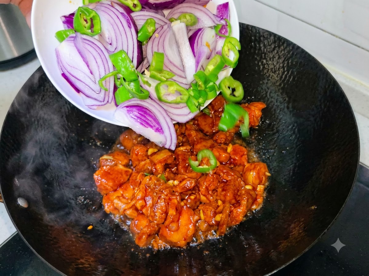 白いお皿から、スライスした紫玉ねぎと緑の唐辛子が、豚バラ肉の炒められている中華鍋に入れられている様子