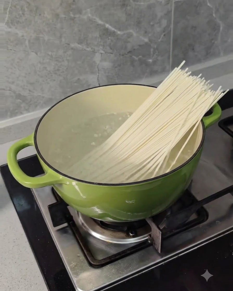 コンロの上の緑色の鍋に入った沸騰したお湯に、乾燥した長い小麦麺が入れられているところ。