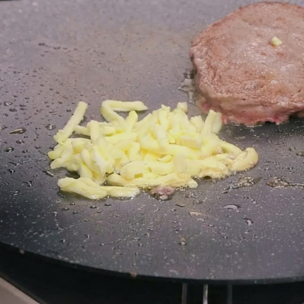 熱したフライパンの上の焼きたてビーフパティの上に、細切りモッツァレラチーズがとろけている様子。