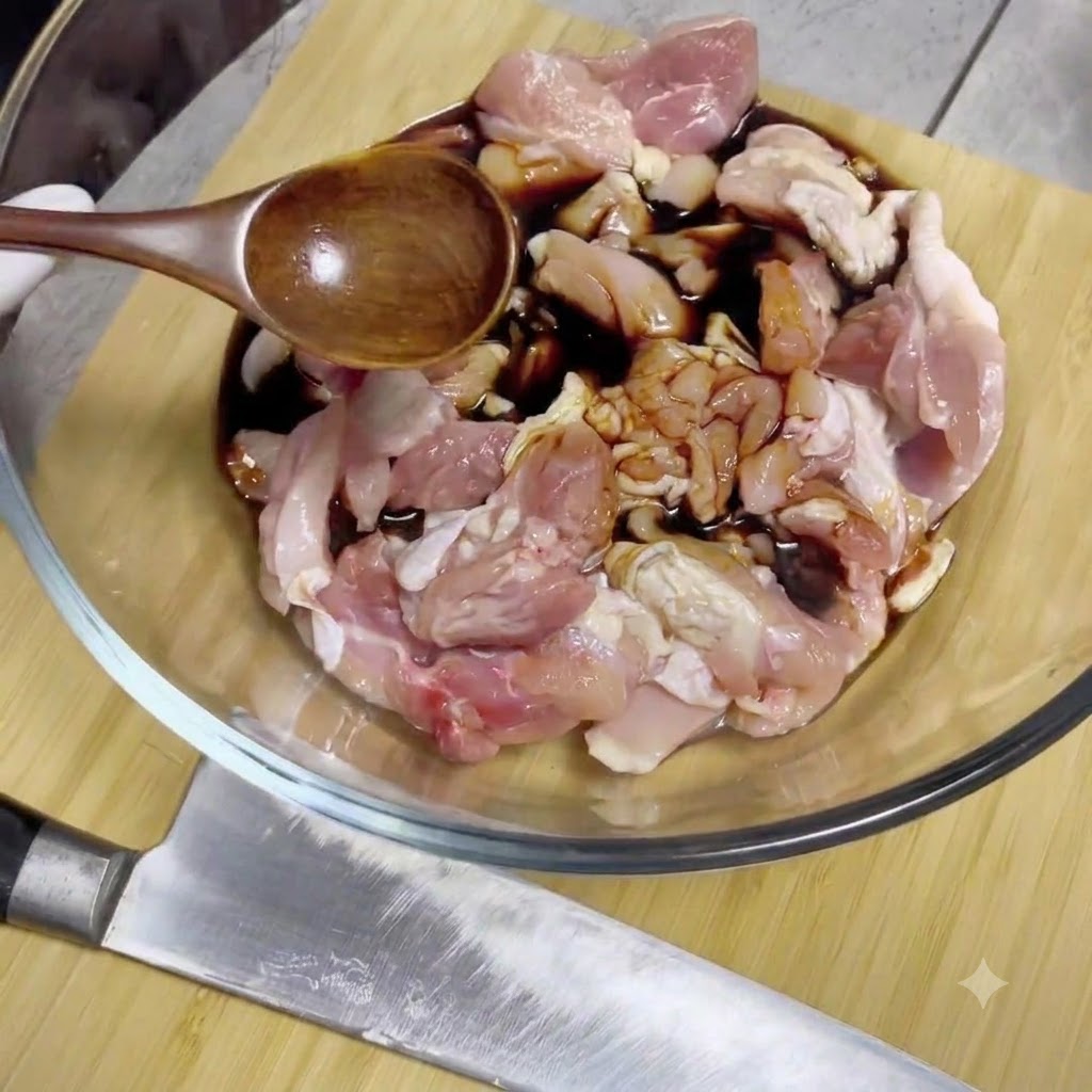 木製のスプーンを使って、ガラスのボウルに入った生の鶏肉の塊に醤油を回しかけている様子。