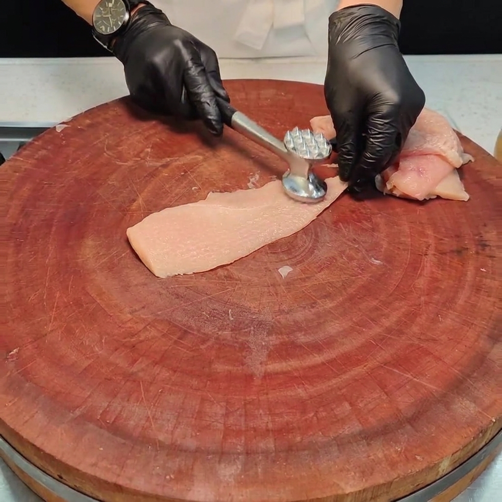 木製ボードの上で、金属製のミートハンマーを使って鶏肉を平らに叩いている様子。