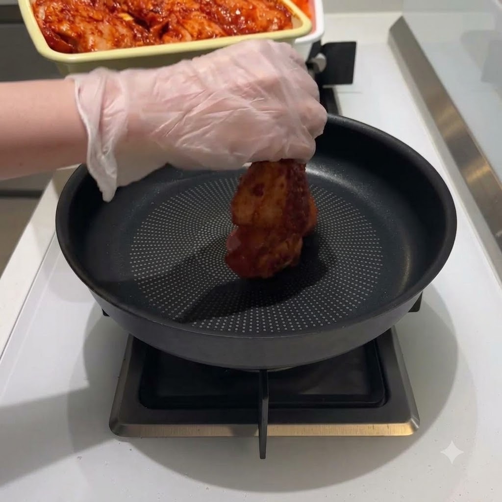 手袋をつけた手が、黒いフライパンの中央に赤い味付け鶏肉の塊を置いている様子。
