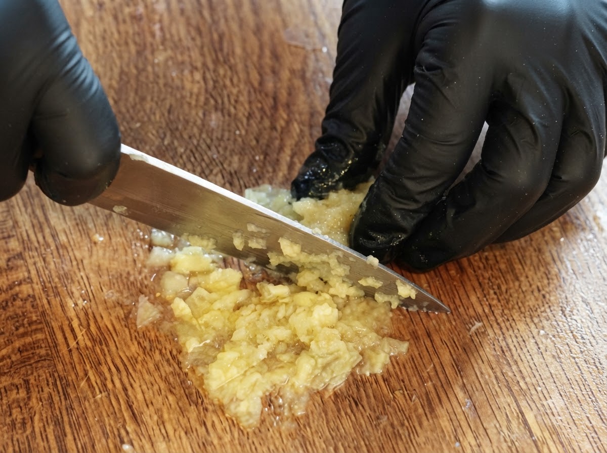 手袋をした両手がナイフを使い、木製のまな板の上で残ったレモンの果肉を刻み、ピューレ状にしているところ。