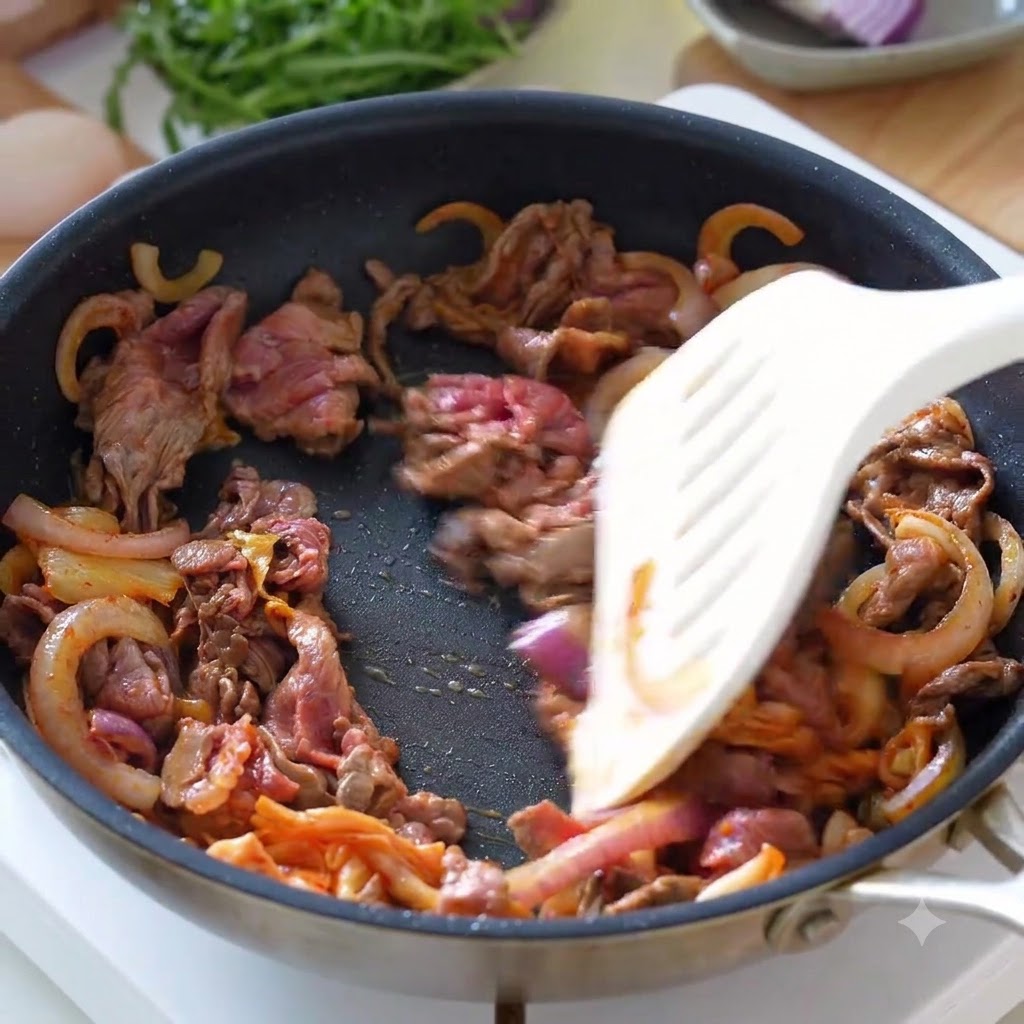 フライパンの中で、薄切り牛肉、玉ねぎ、キムチをヘラで一緒に炒めている様子。