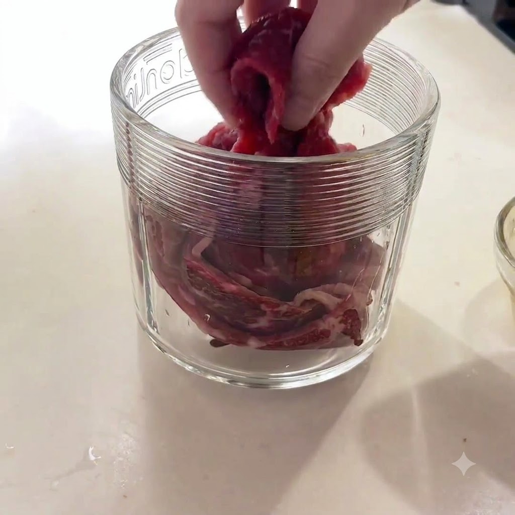 ガラス製のフードプロセッサーの容器に、生の牛肉の塊を入れている手。