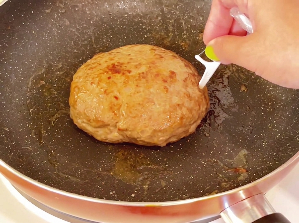 フライパンの中にある厚手の調理済みハンバーグの表面に、爪楊枝が刺されている様子。