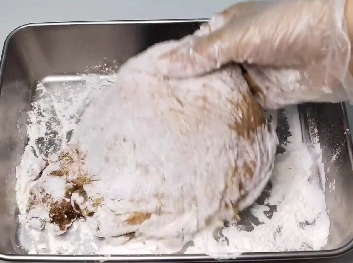 金属製のトレイの上で、手袋をした手が下味をつけた鶏もも肉に白い片栗粉を薄くまぶしている様子。