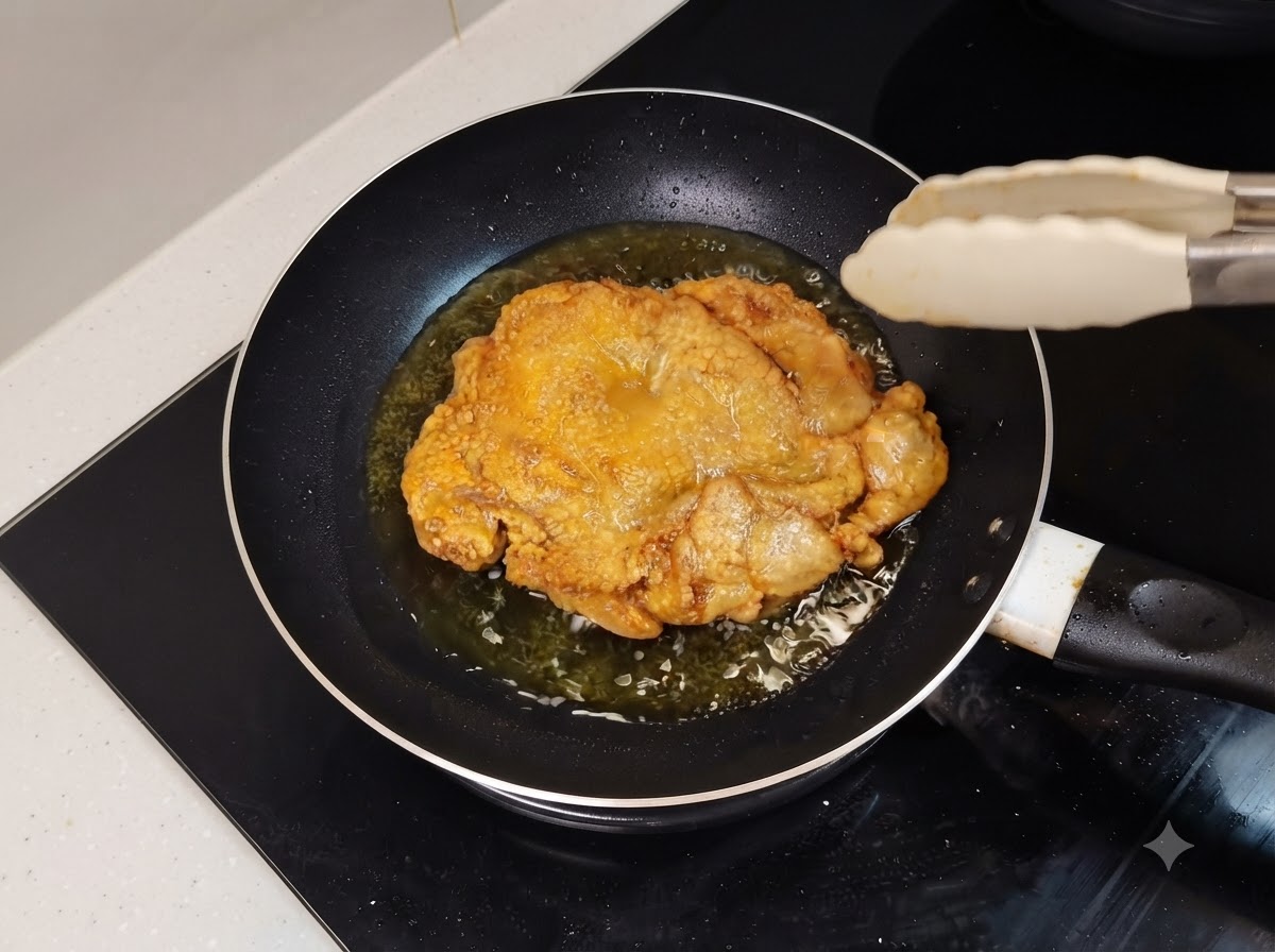 油の入ったフライパンの中で、きつね色に揚がった鶏もも肉が音を立てている様子。近くにキッチントングがある。