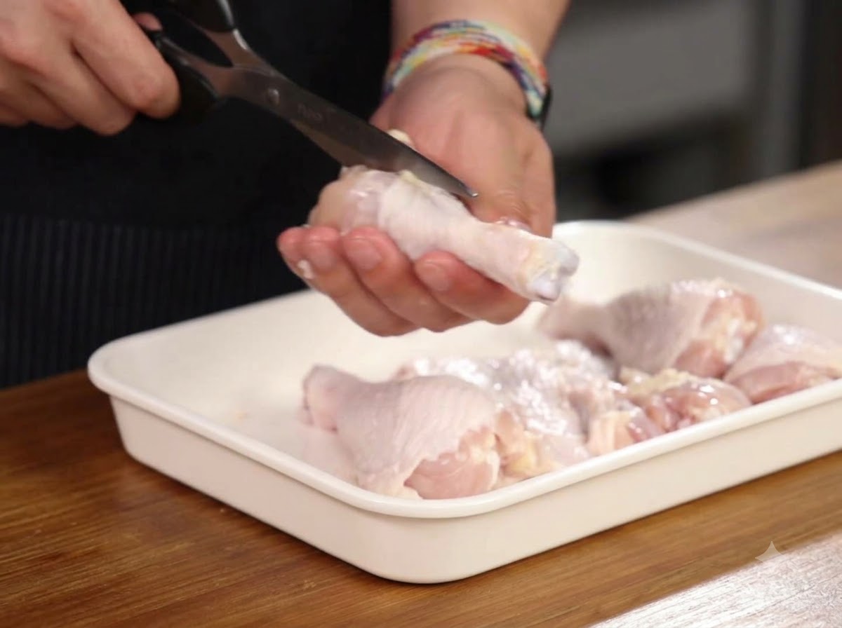 白いトレイの上でキッチンバサミを使い、鶏ドラムスティックの骨に沿って切っている手元。