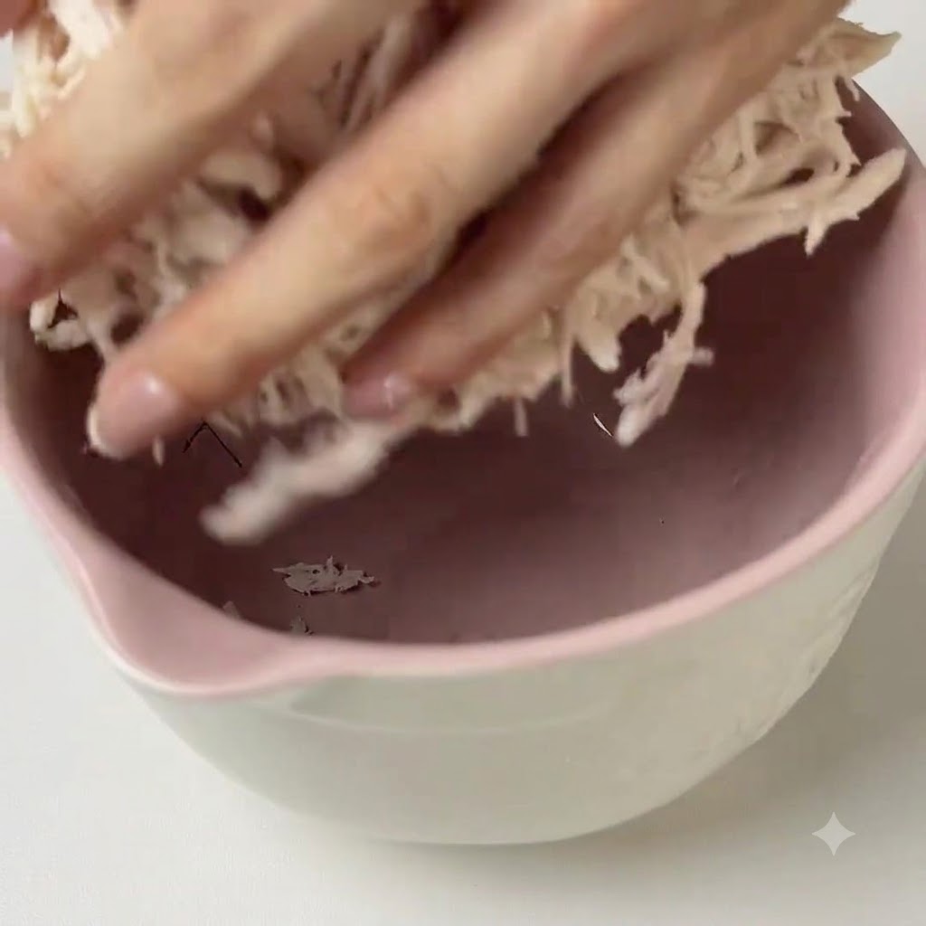 ピンク色のボウルの中で、ほぐした鶏肉に緑色のペストソースを手で混ぜ合わせている様子。